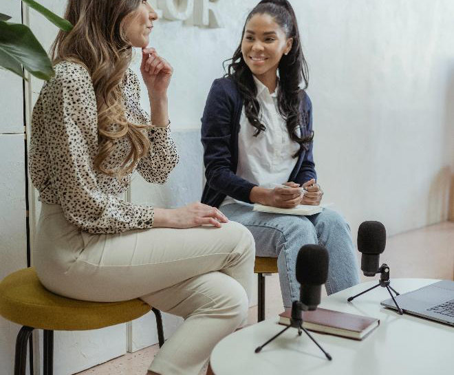 Two women sitting across from each other at a table, each with a microphone in front of them, engaged in a podcast interview