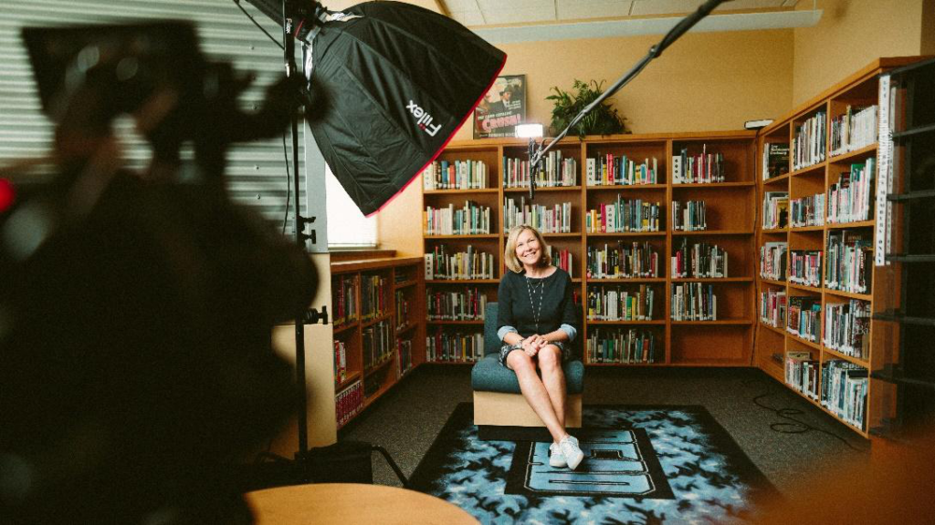 A woman sitting in a chair in a library setting, facing a camera with a softbox light