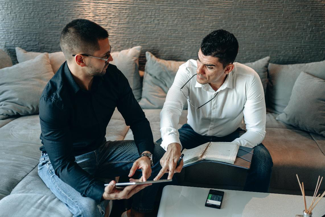 Two men sitting at a table, looking at a tablet screen and discussing content