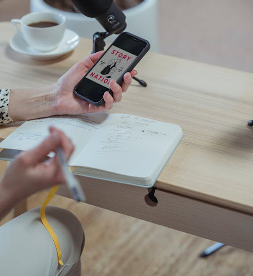 A cropped image of a woman using her smartphone while writing in a diary, seated at a desk.