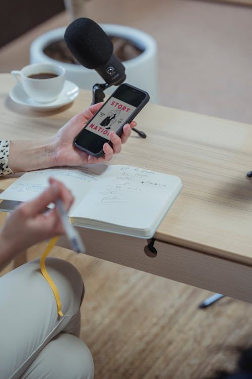 A cropped image of a woman using her smartphone while writing in a diary, seated at a desk.