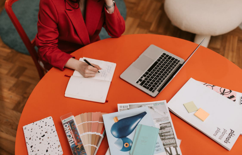 A woman in a red blazer writing in a notebook while seated at a desk, appearing focused and professional.