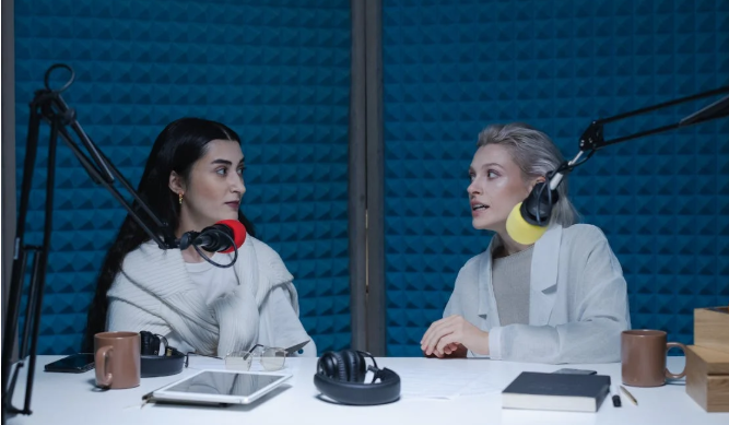 Two women seated in a podcast studio, wearing headphones and engaged in a lively conversation across microphones.